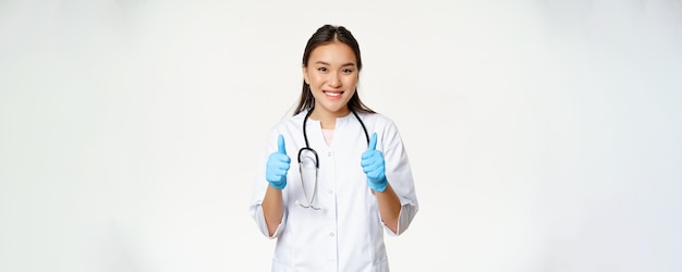 Smiling asian doctor woman in medical uniform and rubber gloves shows thumbs up recommending smth to patient standing over white background