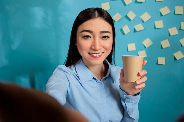 Smiling asian employee holding disposable coffee cup while taking a break from work and talking to friends via video call. Smiling woman at work relaxing before continuing to work on paperwork.