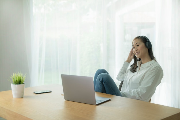 Photo smiling asian female in casual look working with headphones and laptop notebook computer on the desk at home.