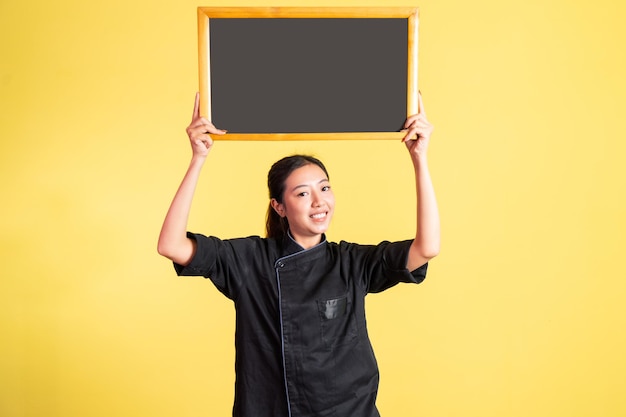 Smiling asian female chef lifting and showing blank blackboard