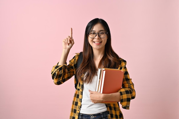 Smiling Asian female college student pointing finger up on empty space on a pink background