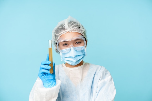 웃고 있는 아시아 여성 의사, 개인 보호 장비를 착용한 간호사, 샘플 테스트 튜브를 보여주고 웃고, 분석하고, 파란색 배경 위에 서 있습니다.