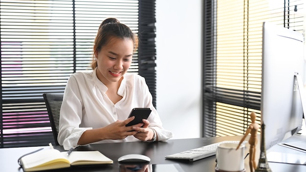 彼女のオフィスの机に座ってスマートフォンを使用して笑顔のアジアの女性従業員