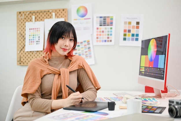 Smiling Asian female graphic designer or interior designer sits at her desk in her studio