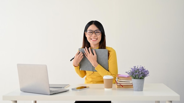 A smiling Asian female holding a book while looking at the camera and sitting at her desk