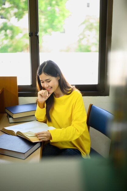 Smiling asian female student reading book doing class assignment in library Education learning and people concept