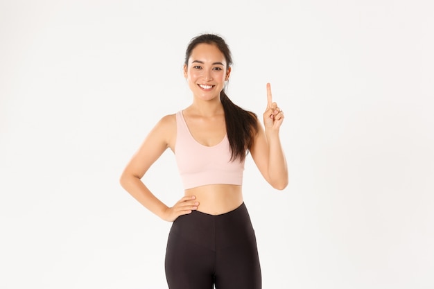 Smiling asian fitness instructor, girl in sportswear teaching lesson, showing rule number one, raise finger and smirk, standing