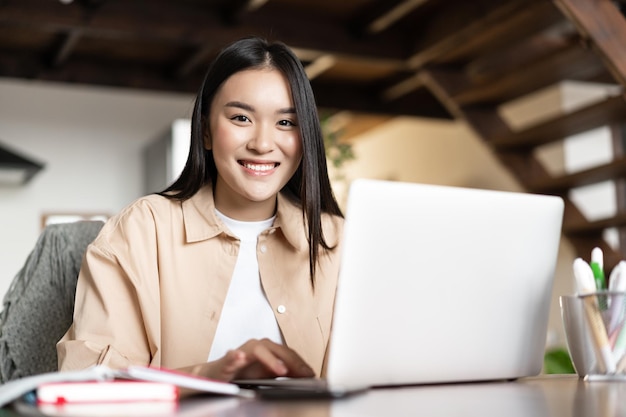 Photo smiling asian girl doing homework studying online on laptop from home young woman working on compute...