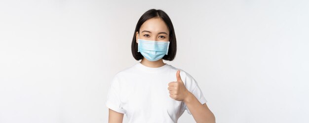 Sorridente ragazza asiatica in maschera medica che mostra i pollici in su approva qualcosa di buono elogio e complimenta la compagnia in piedi su sfondo bianco