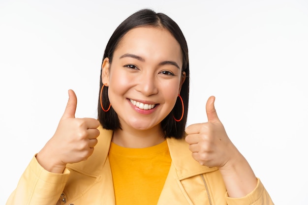 Smiling asian girl showing thumbs up looking pleased approve smth standing over white background