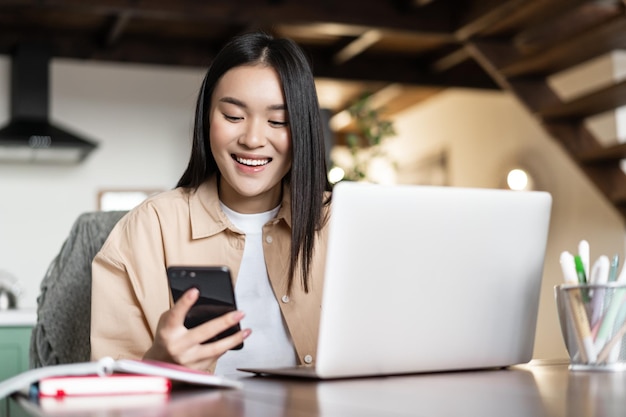 からの仕事のために携帯電話とコンピューターを使用してラップトップの女性の宿題をしている笑顔のアジアの女子学生...