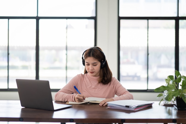 Smiling Asian girl student wears wireless headphones write on the notebook to study language online watch and listen to the lecturer, webinar via video call e-learning at home, distance education