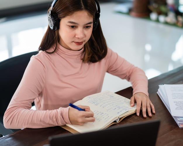 Smiling Asian girl student wears wireless headphones write on the notebook to study language online watch and listen to the lecturer, webinar via video call e-learning at home, distance education