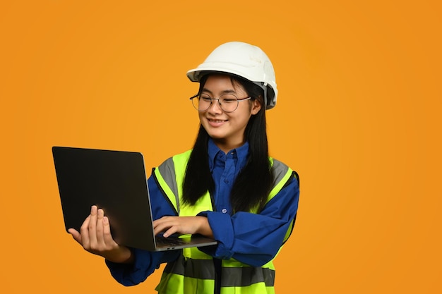 Smiling asian girl wearing helmet holding laptop isolated on yellow background future dream job