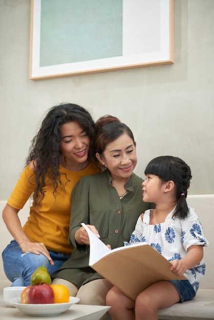 Smiling asian grandmother sitting on sofa with adult daughter and granddaughter and reading book with them