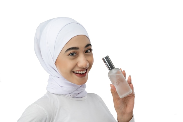 Улыбающаяся азиатская девушка в хиджабе в белом платье держит бутылку сыворотки для ухода за лицом