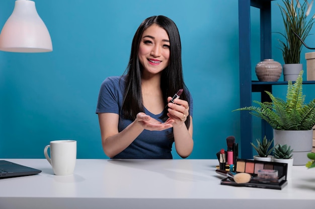 Smiling asian influencer talking with fans presenting makeup lipstick while recording creative tutorial for beauty podcast channel. Social media content creator advertising cosmetic product