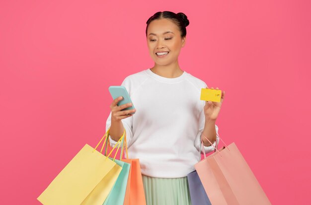 Smiling asian lady holding smartphone credit card and shopping bags