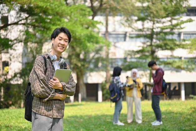 写真 ラップトップとバックパックを持ち、公園に立っている笑顔のアジアの男子大学生