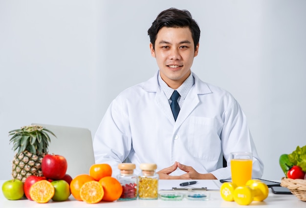 Улыбающийся азиатский диетолог-мужчина сидит за столом с разными свежими фруктами и смотрит в камеру, показывая концепцию здорового питания