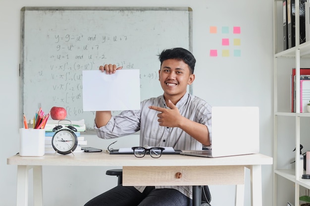 Smiling Asian male teacher sitting on modern workdesk at classroom and showing blank paper with copy