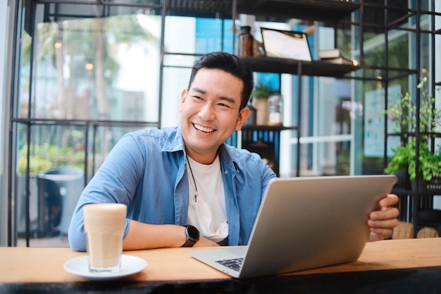 Smiling Asian man in casual shirt using laptop at cafe