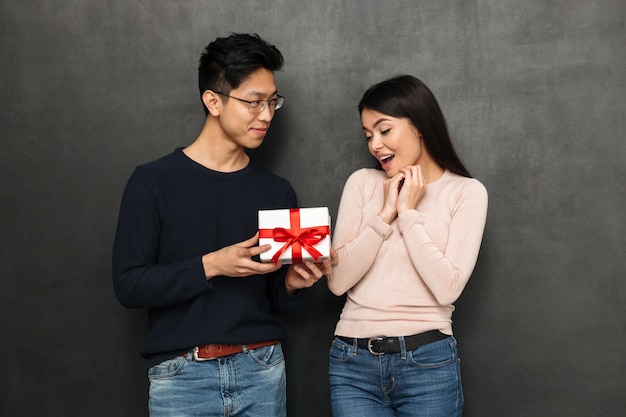 Smiling asian man in eyeglasses presents gift to surprised girlfriend