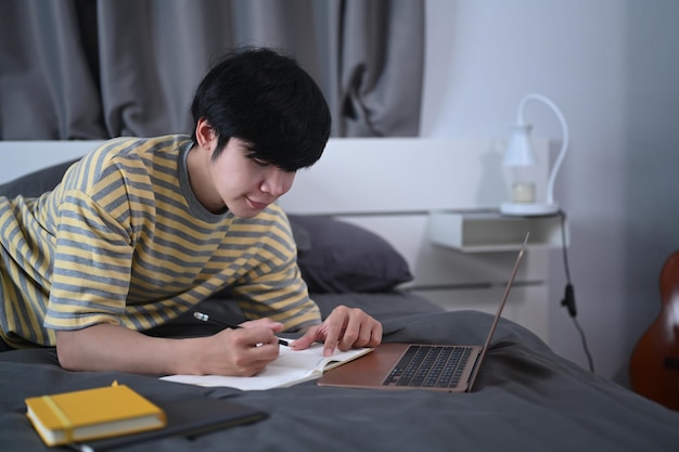 Smiling asian man lying on bed and studying online with laptop computer.