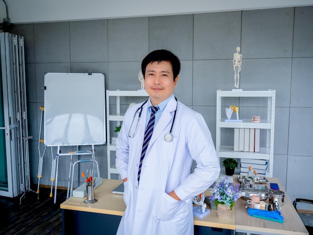 Smiling asian man orthopedic doctor portrait in white coat\
standing near desk bookshelf equipment and tools in medical office\
confident adult male physician or practitioner with\
stethoscope
