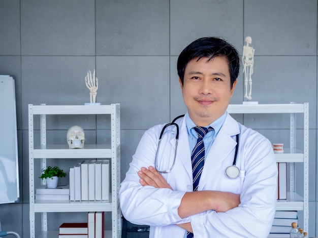 Smiling Asian man orthopedic doctor portrait in white coat standing with crossed arms near bookshelf and equipment in medical office Confident adult male physician or practitioner with stethoscope