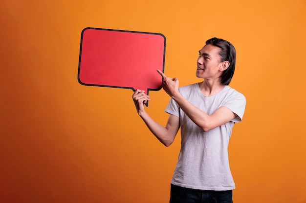 Smiling asian man pointing at empty red speech bubble with copy space, showing advertising mock up. Cheerful teenager holding dialog frame, person with communication cloud