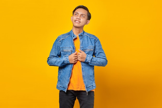 Smiling asian man wearing blue jacket looking at empty space with hope on yellow background