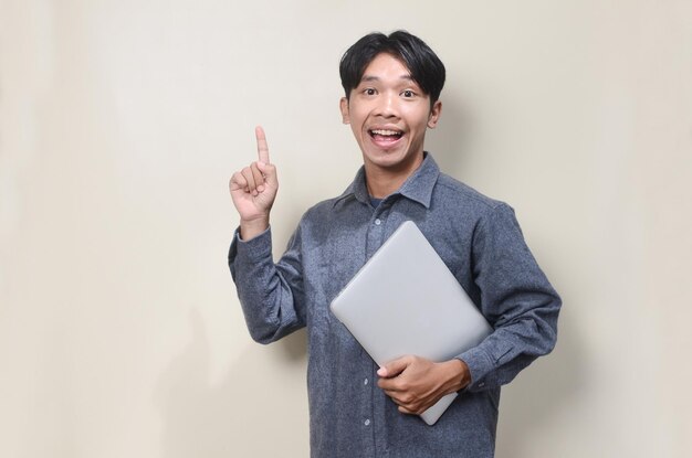 Smiling asian man wearing gray shirt holding laptop computer pointing up on isolated background