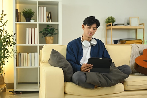 Smiling asian man with headphones browsing internet on digital tablet in living room