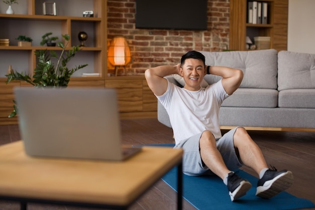 Smiling asian mature man working on abs watching fitness tutorial on laptop and exercising on fitness mat at home