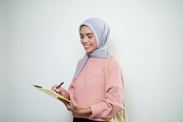 Улыбающаяся азиатка-мусульманка в розовом свитере с сумкой, держащей доску и указывающая на пустую доску