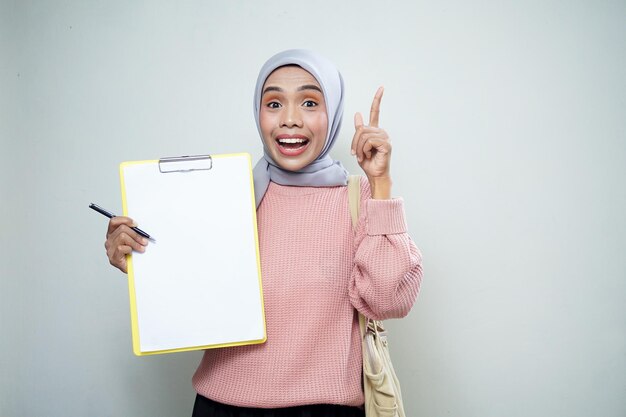 Smiling asian muslim female student in pink sweater with bag holding board and pointing empty board