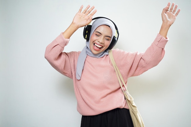 Smiling Asian Muslim female student in pink sweater with bag and listening to music excitedly