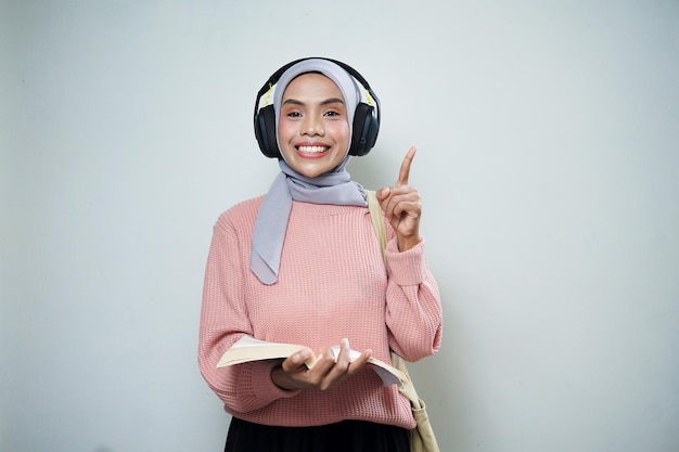 Улыбающаяся азиатская студентка-мусульманка в розовом свитере с сумкой во время прослушивания музыки и чтения книги