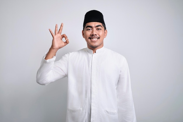 Smiling asian muslim man in white clothes and black skull cap giving okay gesture