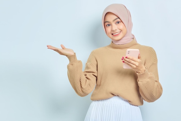 Smiling Asian muslim woman in brown sweater and hijab using a mobile phone showing copy space on hands isolated over white background People religious lifestyle concept