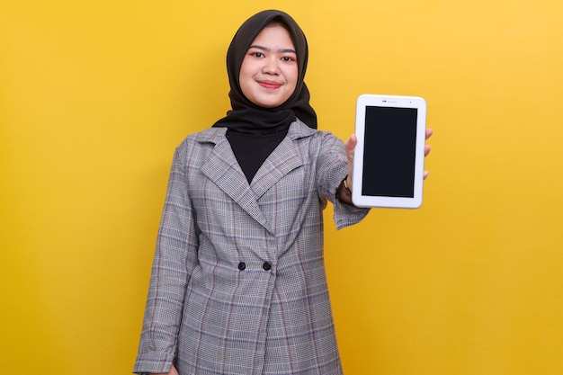 空白のタブレット ph を見せながら黄色の背景に立っているヒジャブを着た笑顔のアジアのイスラム教徒の女性