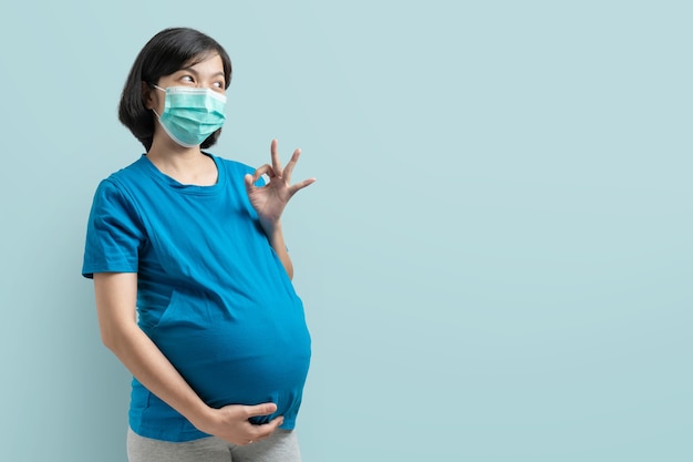 Photo smiling asian pregnant woman with protective face mask and blue t-shirt showing gesture ok