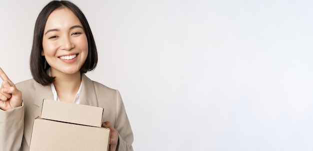 Smiling asian saleswoman pointing finger at copy space holding boxes with delivery order standing ov