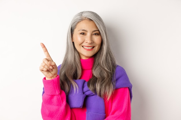 Smiling asian senior woman showing promo offer, looking happy and satisfied while pointing upper left corner, standing over white background.