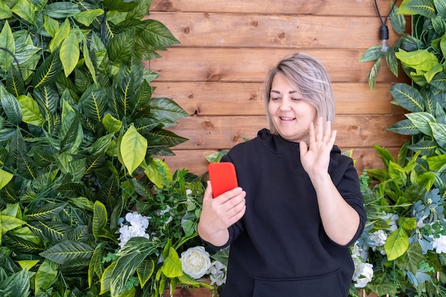 A smiling Asian woman in a black jacket uses a smartphone for online video communication waving her hand greeting friends