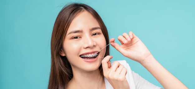 Smiling asian woman cleaning braces on teeth with dental floss\
on blue background, concept oral hygiene and health care.