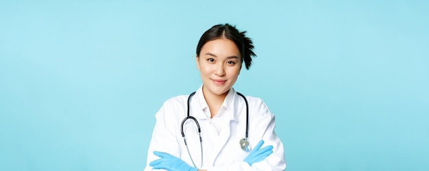 Smiling asian woman doctor nurse in gloves and clinic uniform cross arms and looking determined blue