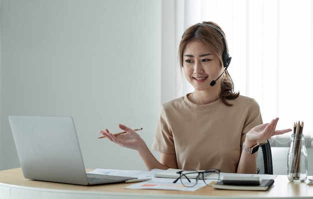 Smiling asian woman freelancer wearing headset, communicating\
with client via video computer call. millennial pleasant\
professional female tutor giving online language class.