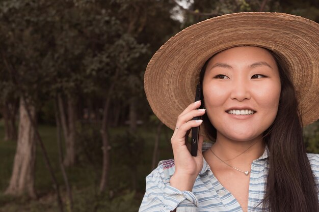 Smiling asian woman in hat talking by phone in city park at summer